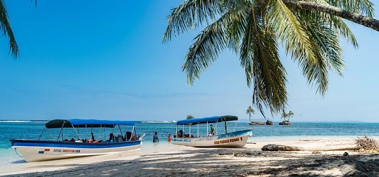 A beautiful, blue-sky day at the beach in Panama with shade from a palm tree and two island-hopping boats resting on shore
