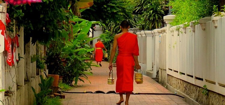 A monk from Laos wearing an orange robe and walking with two buckets in his hands. Laos Facts