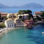 A group of homes along the water in Greece, with green and blue waters and a mountain in the background.