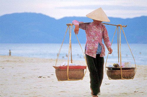 a woman wearing a triangular hat carrying baskets on a stick over her shoulder