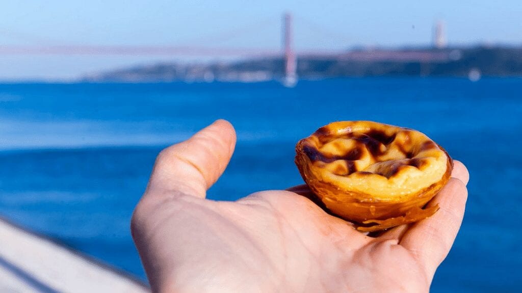 a hand holding a small pastry in the background is the blue sea and a suspension bridge