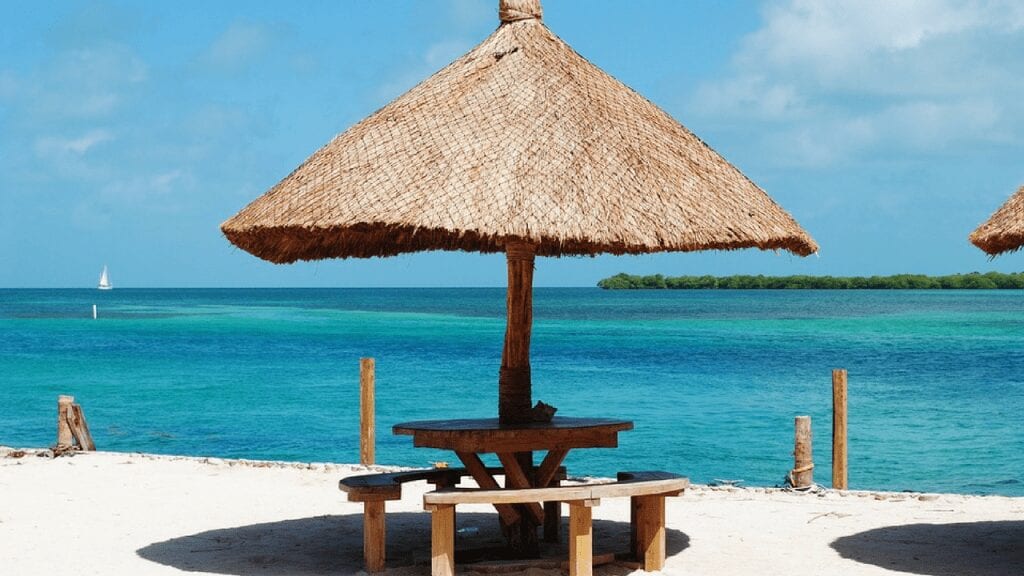 Ambergris Caye, Belize, white sand beach with wooden table and leaf umbrella. One of the Cheapest Places to Live in the Caribbean According to Forbes 2019