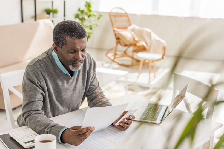 Senior man looking at utility bill while sitting near laptop