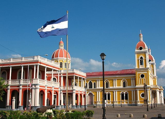 Old colonial buildings of orange and yellow with the Nicaragua flag waving in the air and a cannon out front.