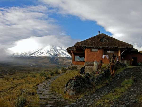 a small orange stone house with llamas. A view of a mountaine at the distance with the tip covered with clouds and a blue sky 