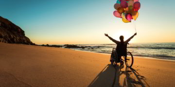 A person on a wheelchair with colorful balloons at the beach. disability friendly countries