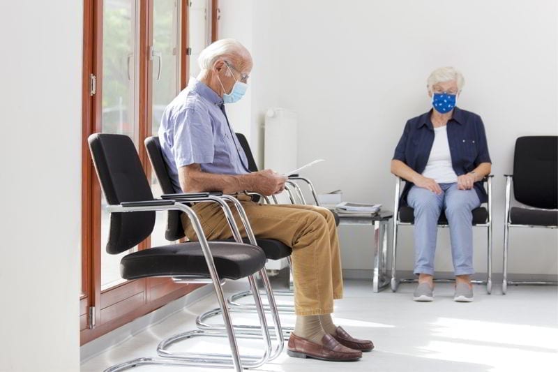 Senior woman and man sitting with face masks in a bright waiting room of a hospital or an office. 