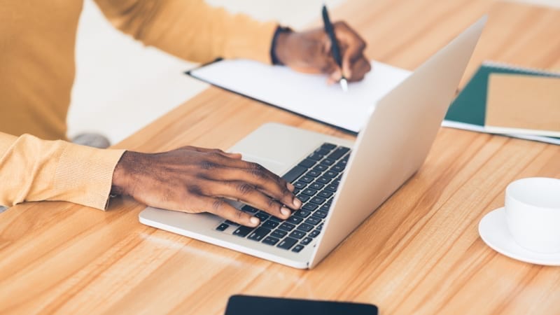 Close up businessman writing content on notebook, using laptop at wooden table at office