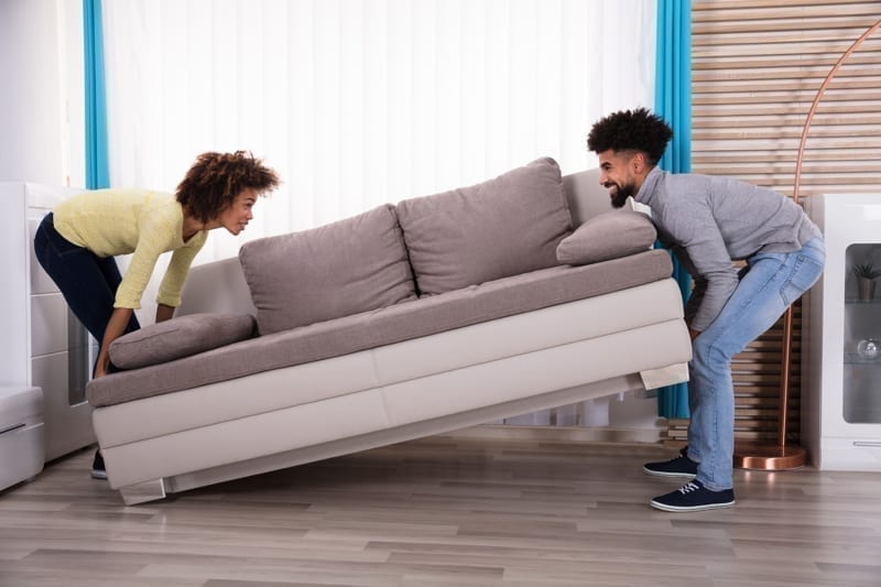 Side View Of Smiling Couple Lifting Sofa In Living Room. 