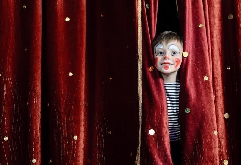 Young Boy Wearing Clown Make Up Peering Out Through Opening in Red Stage Curtains. 