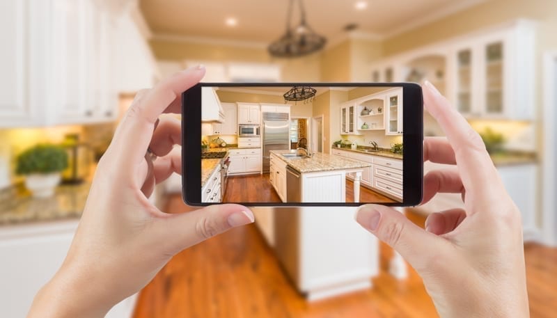 Hands Holding Smart Phone Displaying Photo of Kitchen Behind.