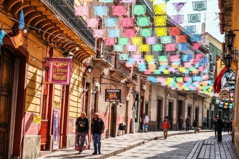 Mexicans walking on cobblestone street with sun lit colorful banner flags hanging above, Guanajuato Mexico