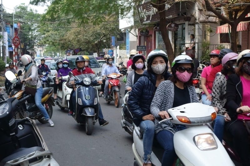 Scooters with riders, some wearing face masks, on a busy street in Hanoi, Vietnam