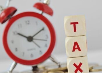 The word Tax written with wooden blocks and alarm clock on coins in the background.
