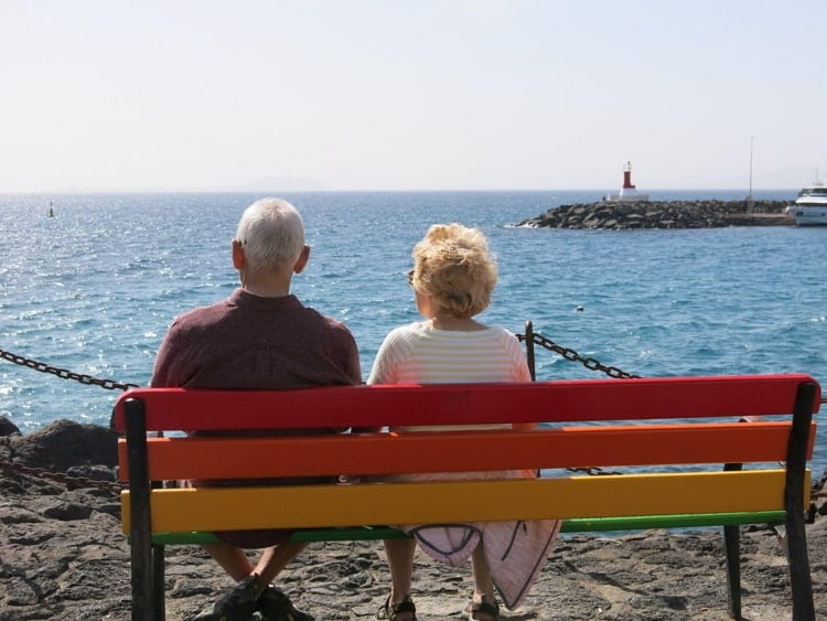 Rear view of an elderly couple sitting on a multi-coloured bench, enjoying some winter sunshine abroad & looking out to sea