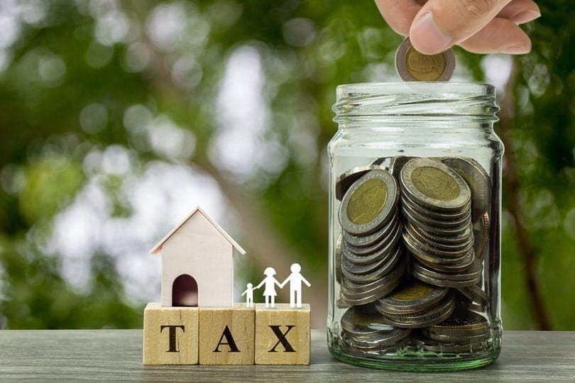 Hand holding coin over stack coins in a jar and small residential house model with family on wooden block on wood