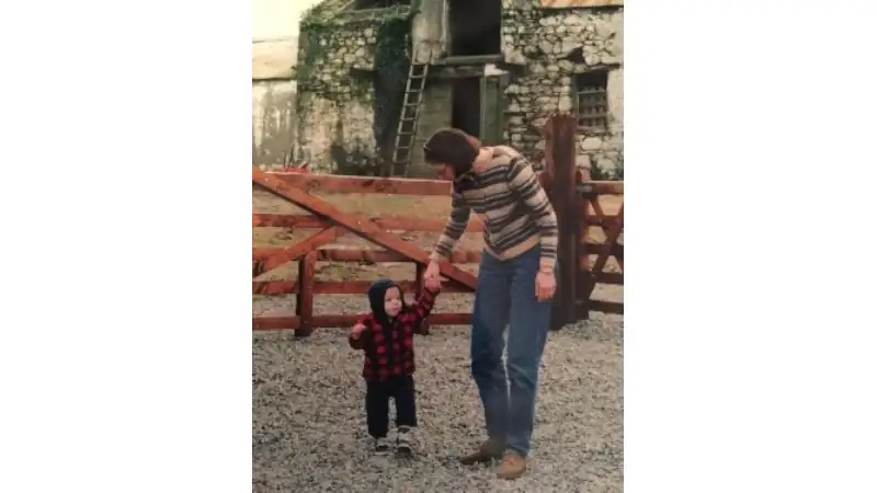 Adult holding hands with a small child in a rustic courtyard with stone buildings and wooden gates in the background.