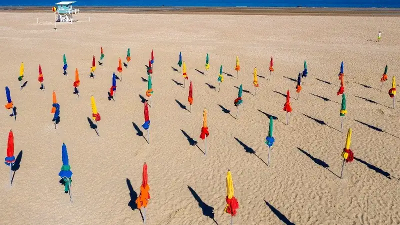 Aerial view of colorful closed beach umbrellas spread across a wide sandy beach beside the ocean.