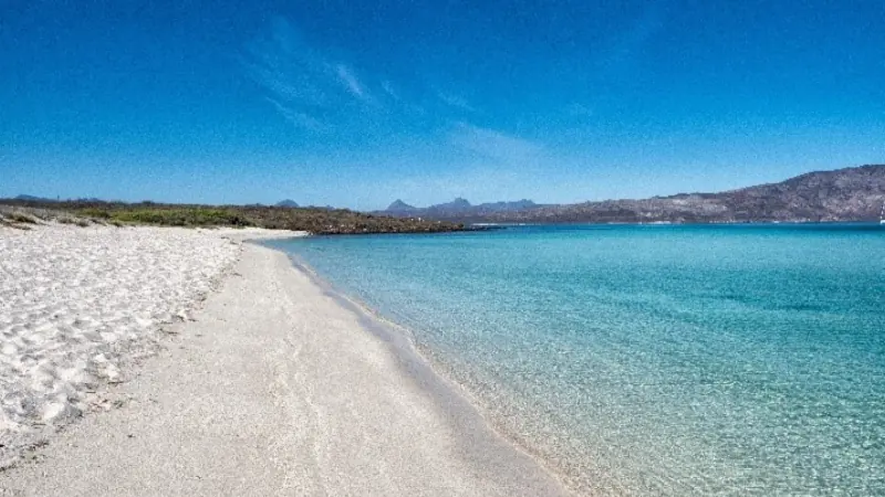 Pristine beach with white sand and clear turquoise water in Loreto Bay National Marine Park, Mexico, under a bright blue sky.