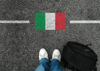 A man with a shoes and backpack is standing on asphalt next to flag of italy