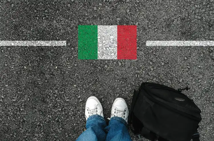 A man with a shoes and backpack is standing on asphalt next to flag of italy