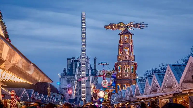 Festive Christmas market scene with brightly lit stalls, a tall ferris wheel, and decorative holiday towers under an evening sky.