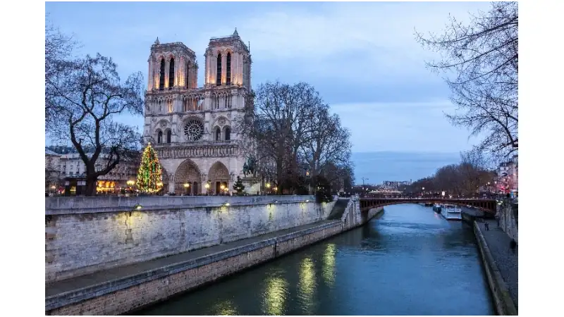 Notre-Dame Cathedral overlooking the Seine at dusk with a lit Christmas tree by the riverbank.