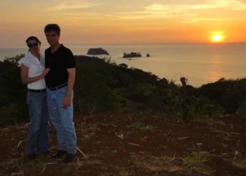 Couple standing on a hilltop at sunset with an ocean view and small islands in the background.