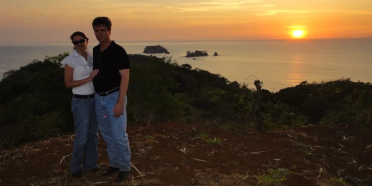 Couple standing on a hilltop at sunset with an ocean view and small islands in the background.