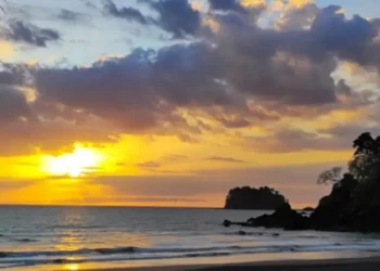 A golden sunset over the beach at Los Islotes, with gentle waves, dramatic clouds illuminated by the fading light, and rocky coastal outcrops silhouetted against the horizon.