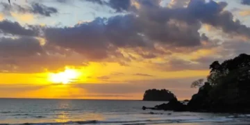 A golden sunset over the beach at Los Islotes, with gentle waves, dramatic clouds illuminated by the fading light, and rocky coastal outcrops silhouetted against the horizon.