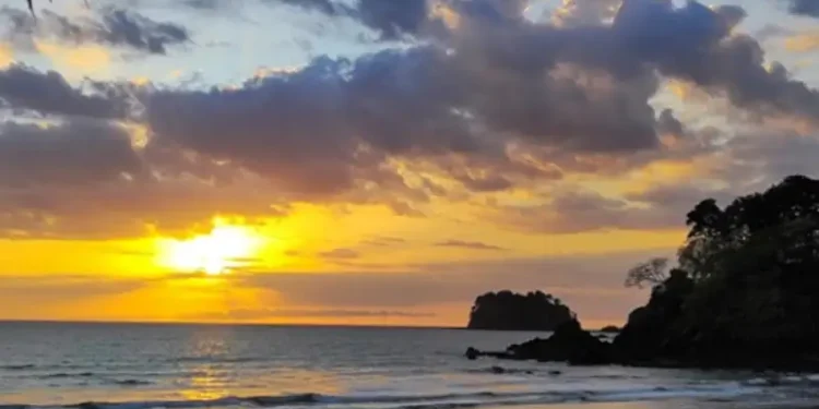 A golden sunset over the beach at Los Islotes, with gentle waves, dramatic clouds illuminated by the fading light, and rocky coastal outcrops silhouetted against the horizon.