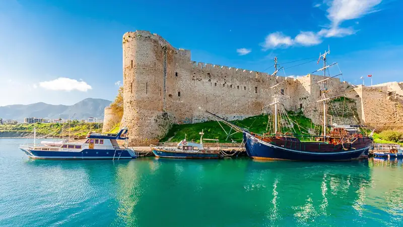 scenic view of Kyrenia Harbour in Northern Cyprus, showing a medieval stone castle overlooking turquoise water, with traditional boats moored along the harbor and mountains in the background.