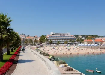 Beachfront promenade in Cascais, Portugal, with palm trees, people on the sandy beach, and calm blue water under a clear sky