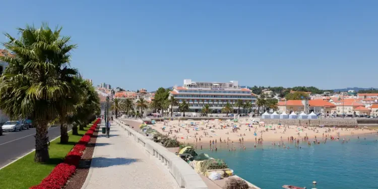 Beachfront promenade in Cascais, Portugal, with palm trees, people on the sandy beach, and calm blue water under a clear sky