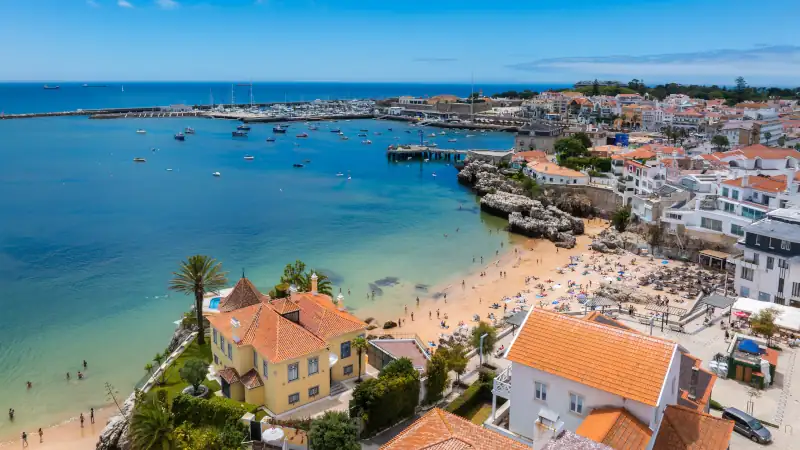 Aerial view of Cascais, Portugal, showing a small beach, colorful buildings with red-tiled roofs, a marina with boats, and clear blue coastal waters