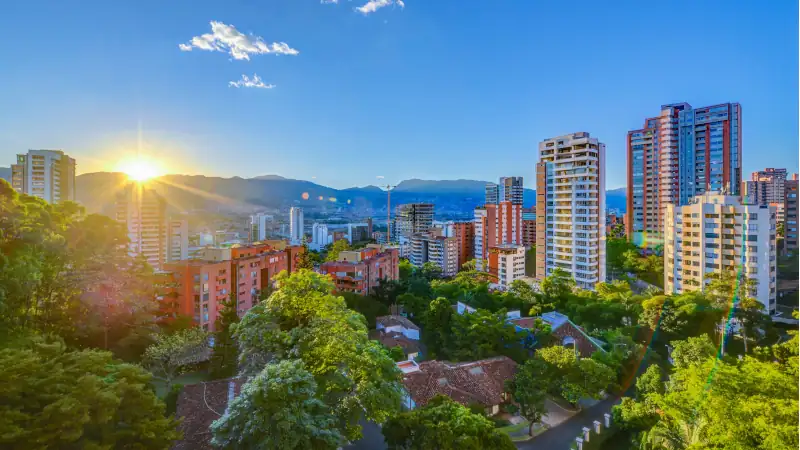 Cityscape of Medellín, Colombia at sunset, with modern high-rise buildings, lush greenery, and mountains in the background under a clear blue sky
