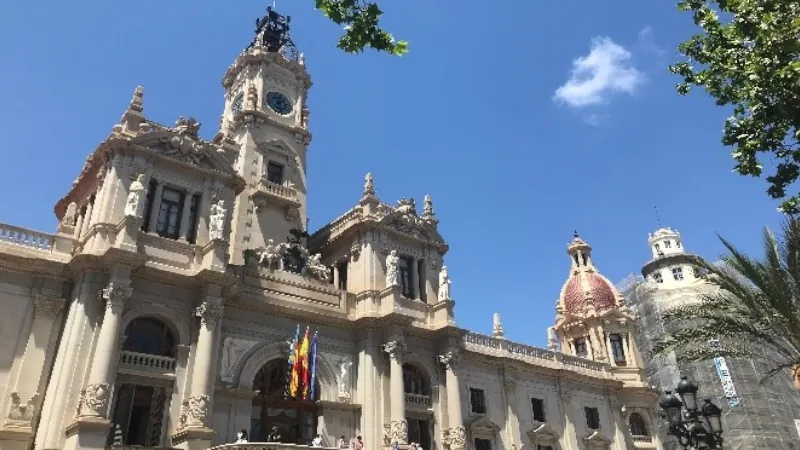 Historic building in Valencia, Spain, featuring ornate architecture, a clock tower, and domed rooftops under a sunny blue sky with tree branches framing the scene