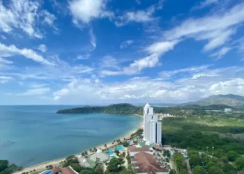 A coastal resort with a beachfront hotel, turquoise ocean views, and lush green hills under a bright blue sky.