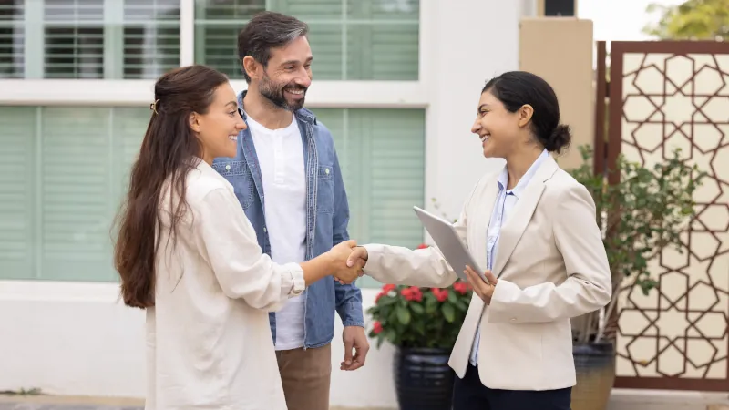 Happy couple of customers and young realtor woman shaking hands outside of house. New homeowners thanking real estate agent for help with mortgage approval, giving handshake