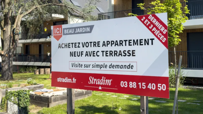 Strasbourg, France - May 12, 2025: A large real estate sign advertises new apartments with terraces by Stradim in front of a modern wood-paneled residential building under a clear blue sky in France