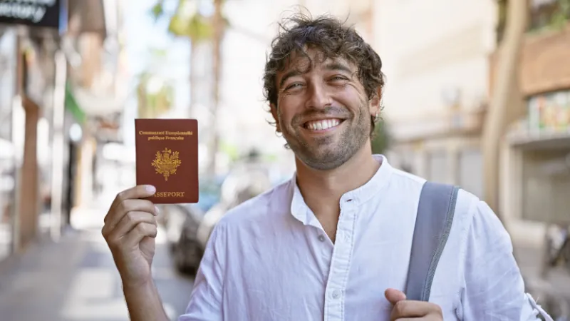 bearded hispanic man with green eyes holds french passport on a sunny urban street.