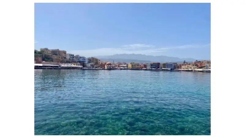 A wide coastal view shows clear turquoise water in the foreground with a waterfront town stretching along the shoreline.