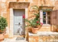 Sunlit rustic wall with a blue door, shuttered window, and potted plants.
