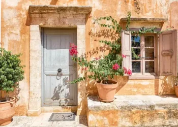 Sunlit rustic wall with a blue door, shuttered window, and potted plants.