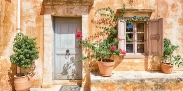 Sunlit rustic wall with a blue door, shuttered window, and potted plants.