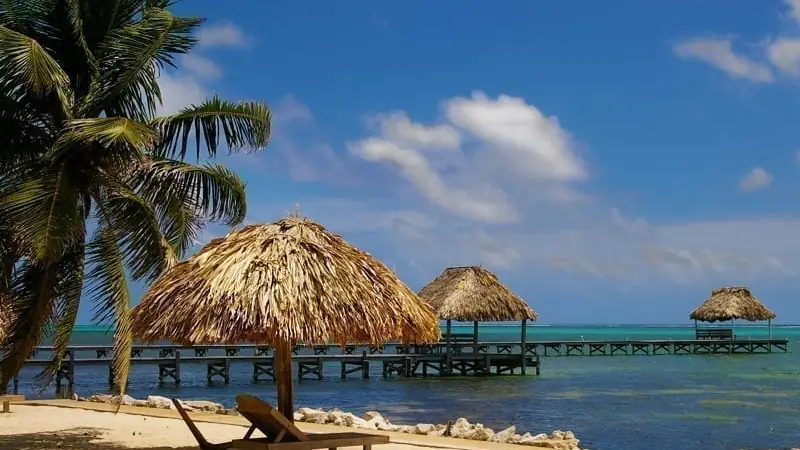 Beach with palm trees, thatched umbrellas, lounge chairs, and wooden piers extending over turquoise water.