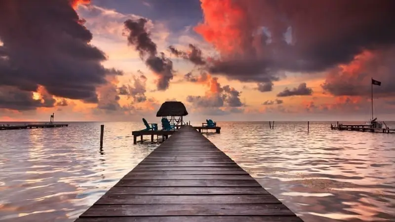 Wooden pier leading to a thatched hut over calm water at sunset, with dramatic clouds and colorful sky.