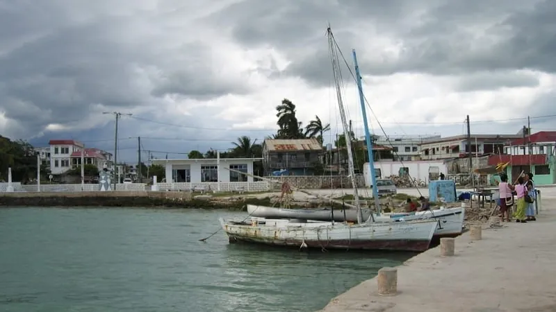 Small sailboat docked at a seaside town with overcast skies and people gathered near the shore.