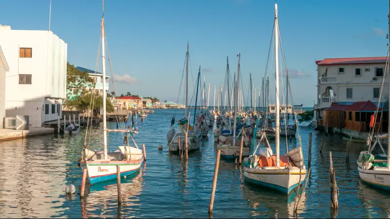 Harbor in Belize with sailboats docked along calm water, surrounded by colorful buildings under a clear blue sky.